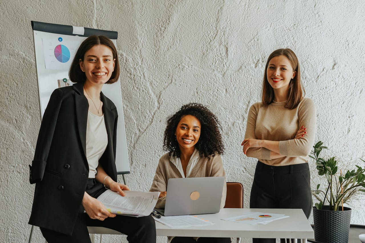 Tres mujeres ejecutivas posando en una oficina junto a una mesa con un portátil y una pizarra.